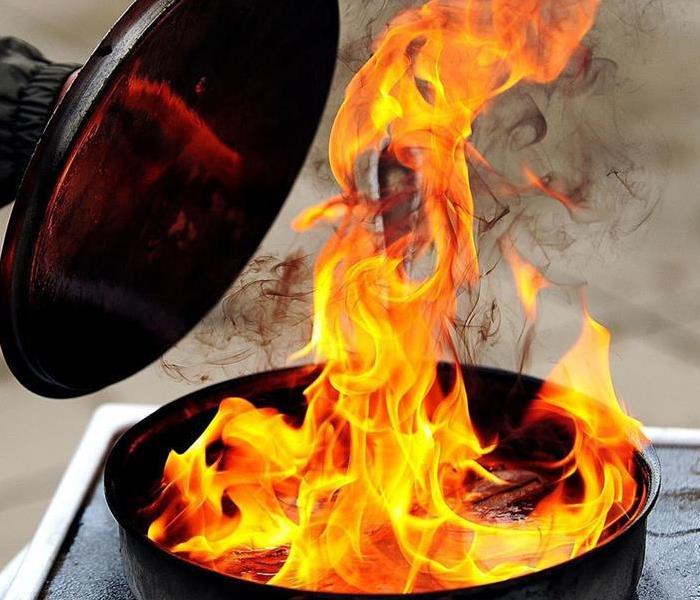 A fire in a pan on the stove with an arm attempting to put a lid on it.