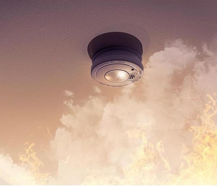 A smoke detector on the ceiling with smoke and flames rising towards it.